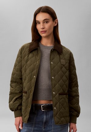 Donna con giacca trapuntata verde oliva con colletto e polsini marroni sopra un maglione corto grigio e jeans blu, in piedi su sfondo grigio.