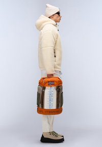 Veste en polaire beige avec finition texturée, pantalon crème, bottes de randonnée marron. Sac à dos orange et kaki avec détails de logo et de drapeau. Les accessoires comprennent un bonnet beige et des lunettes de soleil.