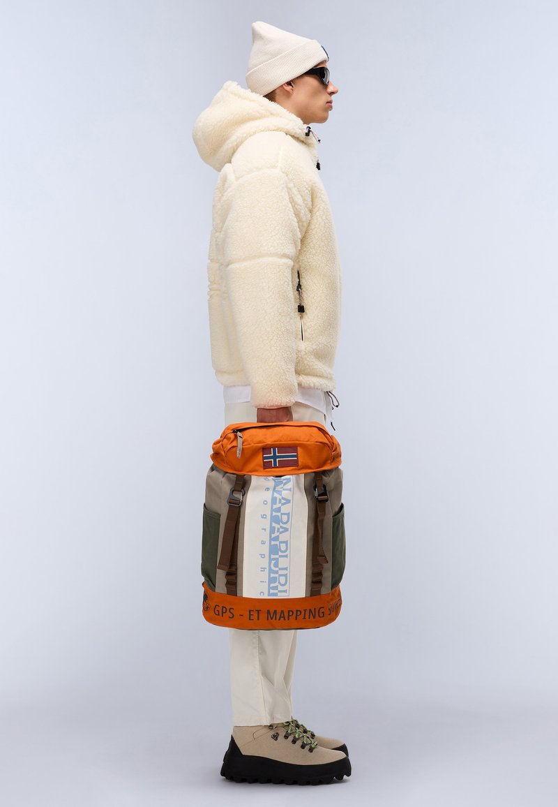 Veste en polaire beige avec finition texturée, pantalon crème, bottes de randonnée marron. Sac à dos orange et kaki avec détails de logo et de drapeau. Les accessoires comprennent un bonnet beige et des lunettes de soleil.