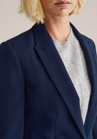 Blonde persona, kas valkā tumši zilu žaketi virs gaiši pelēka aukliņu džemperī, redzama no pleciem līdz vidus krūtīm uz neitrāla fona.