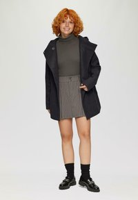 Dunkelgrauer Mantel mit strukturiertem Design, über einem grünen Rollkragenpullover, kombiniert mit einem grauen gestreiften Rock, schwarzen Socken und glänzenden schwarzen Schuhen.