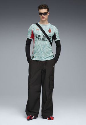 Camiseta deportiva Puma en azul claro con acentos en negro y rojo, con un patrón y texto, combinada con pantalones cargo negros y zapatos con rayas rojas.