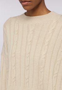 Maglione lavorato a maglia beige con texture a coste e motivo a trecce. Collo rotondo e vestibilità ampia con spalle scese. Tessuto morbido.