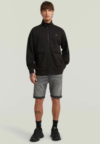 Veste noire zippée avec deux grandes poches avant, portée sur un short en denim gris et des baskets noires, avec une coupe décontractée et un col haut.