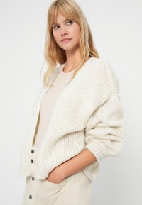 Femme blonde portant un cardigan blanc cassé doux et oversize par-dessus un haut beige clair et une jupe crème boutonnée, mains dans les poches, regardant sur le côté.