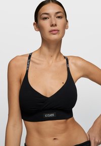 Bralette negro con diseño cruzado, cuenta con tirantes ajustables con detalles de logo. Textura suave de algodón y silueta ajustada.