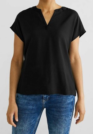 Person wearing a black short-sleeve V-neck blouse paired with blue denim jeans, standing against a plain light background.