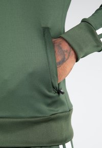 Grüner Sportpullover mit Reißverschlusstasche, gerippten Bündchen und Saum. Glatte Stofftextur mit weißen Streifenakzenten an den Ärmeln.
