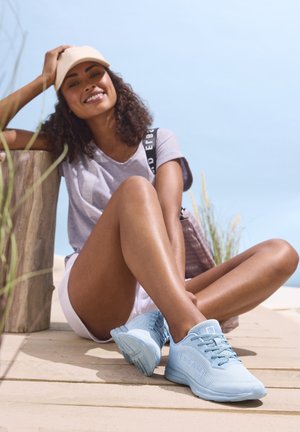 Light blue athletic shoes with textured fabric, breathable design, and laces. Worn by a person seated on wooden decking, dressed casually.