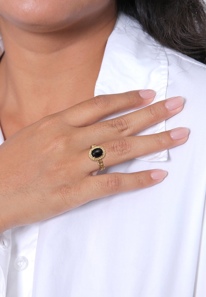 Main avec des ongles vernis rose clair portant une bague en or avec une pierre ovale noire reposant sur le col d'une chemise blanche.