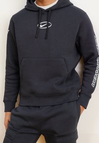 Mörkgrå sweatshirt med dragsko och ficka på magen; har en vit logotyp på bröstet och text på högerärm. Slät textur.