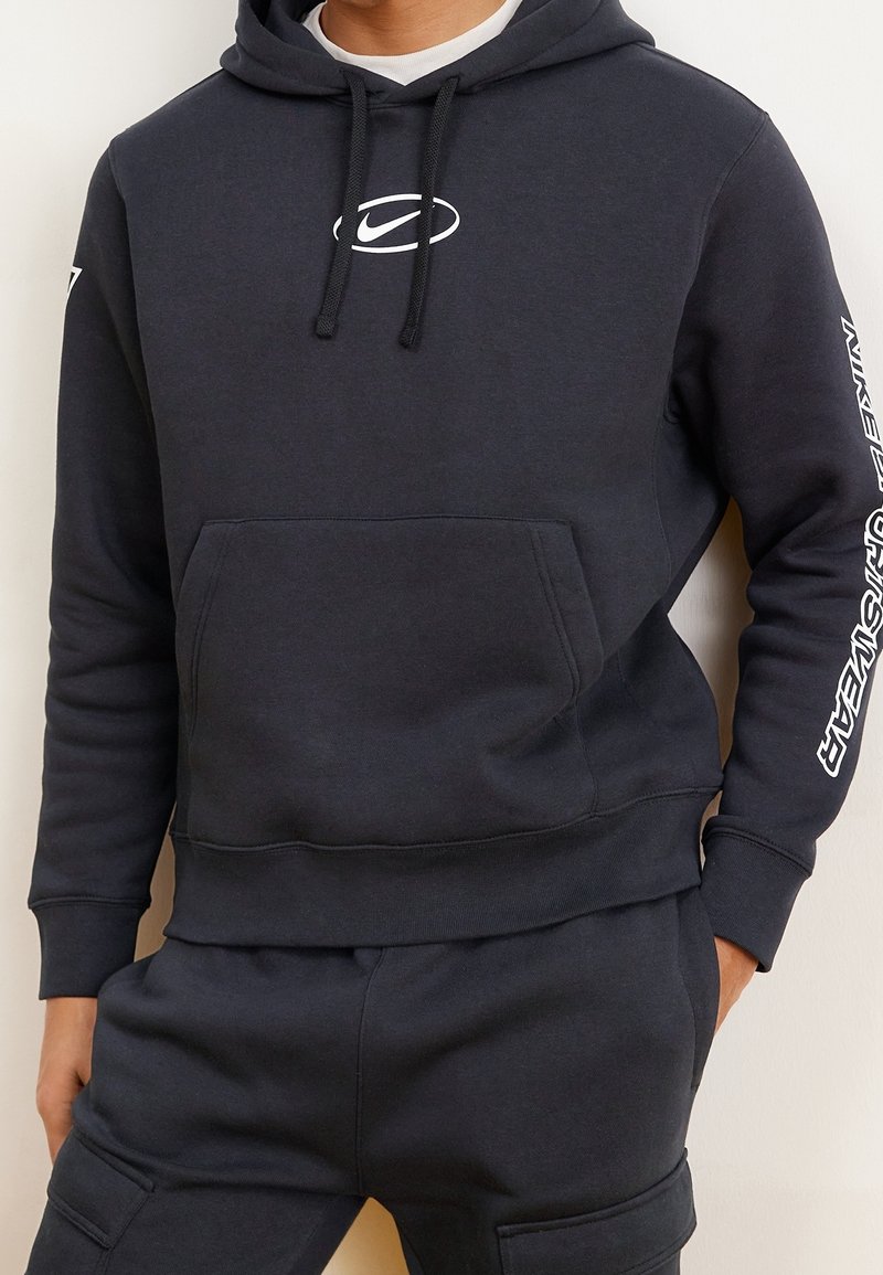 Mörkgrå sweatshirt med dragsko och ficka på magen; har en vit logotyp på bröstet och text på högerärm. Slät textur.