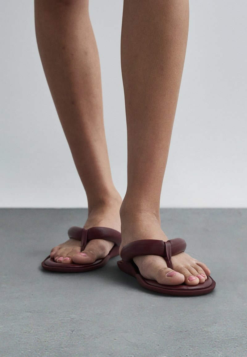 Bare feet with pink nail polish wearing dark red padded thong sandals standing on gray floor against plain light wall.