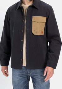 Sort skjortejakke med en tan corduroy brystlomme, knap-front lukning og lange ærmer. Stoffet virker holdbart med en tekstureret finish.