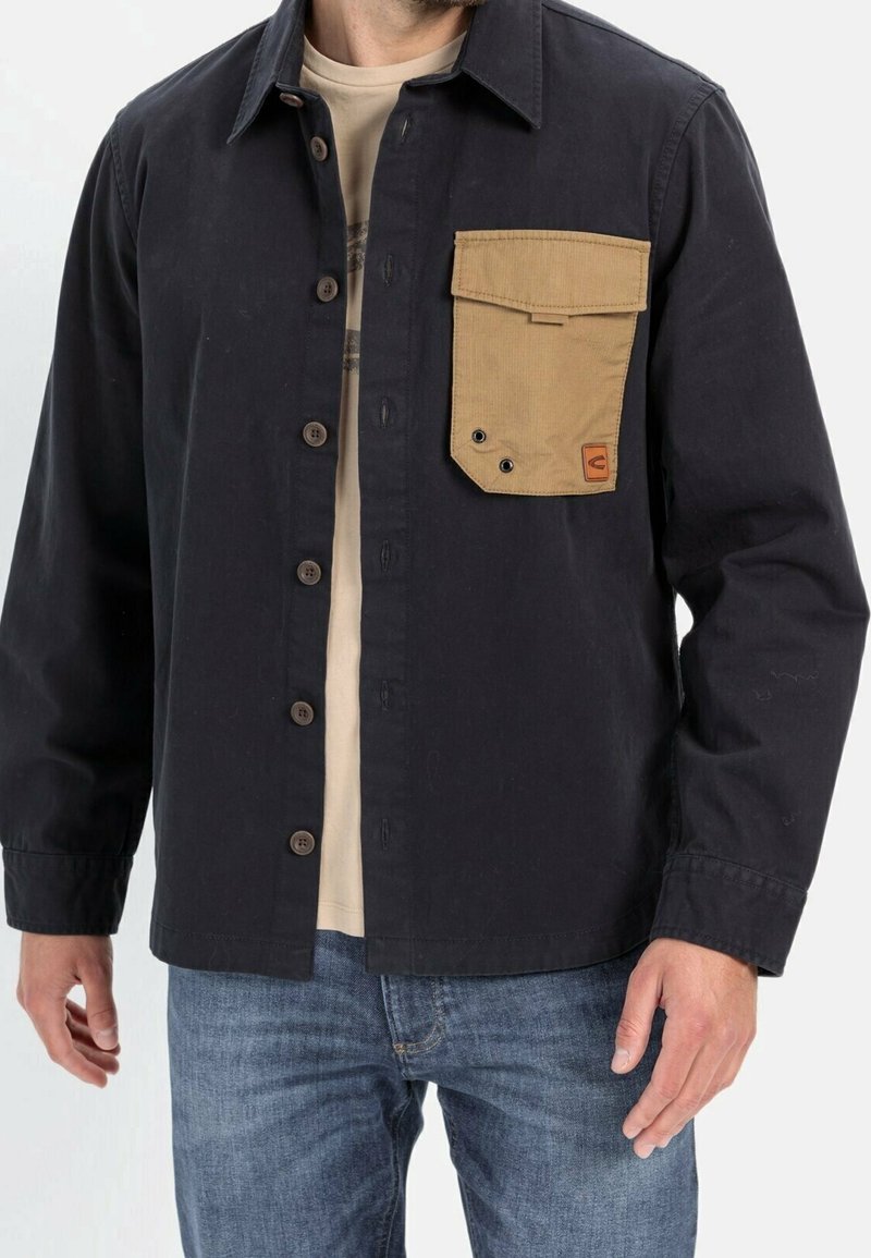 Sort skjortejakke med en tan corduroy brystlomme, knap-front lukning og lange ærmer. Stoffet virker holdbart med en tekstureret finish.