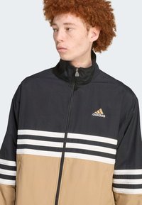 Črna in peščena vetrovka z belimi in črnimi črtami. Ima zadrgo spredaj in vezeno zlato Adidasovo logo na prsih.