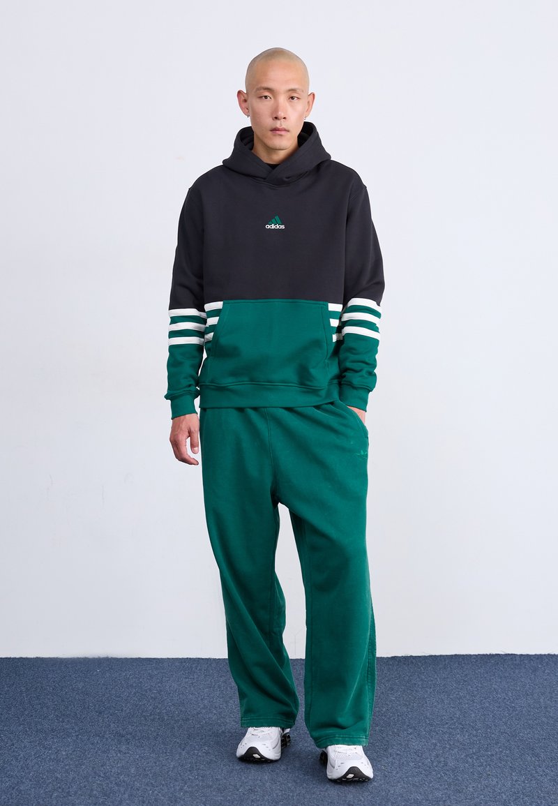 Hombre de pie con sudadera negra y verde de Adidas y pantalones de chándal verdes con zapatillas blancas, contra un fondo blanco liso.