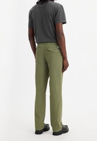 Olivgrön byxor i bomullstwill, raka ben, med två bakfickor och kontrasterande svarta skor. Grå T-shirt synlig.