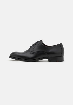 Chaussure de ville en cuir noir avec un bout rond, cinq œillets et un design minimaliste. Elle présente un talon plat et des détails de couture subtils.