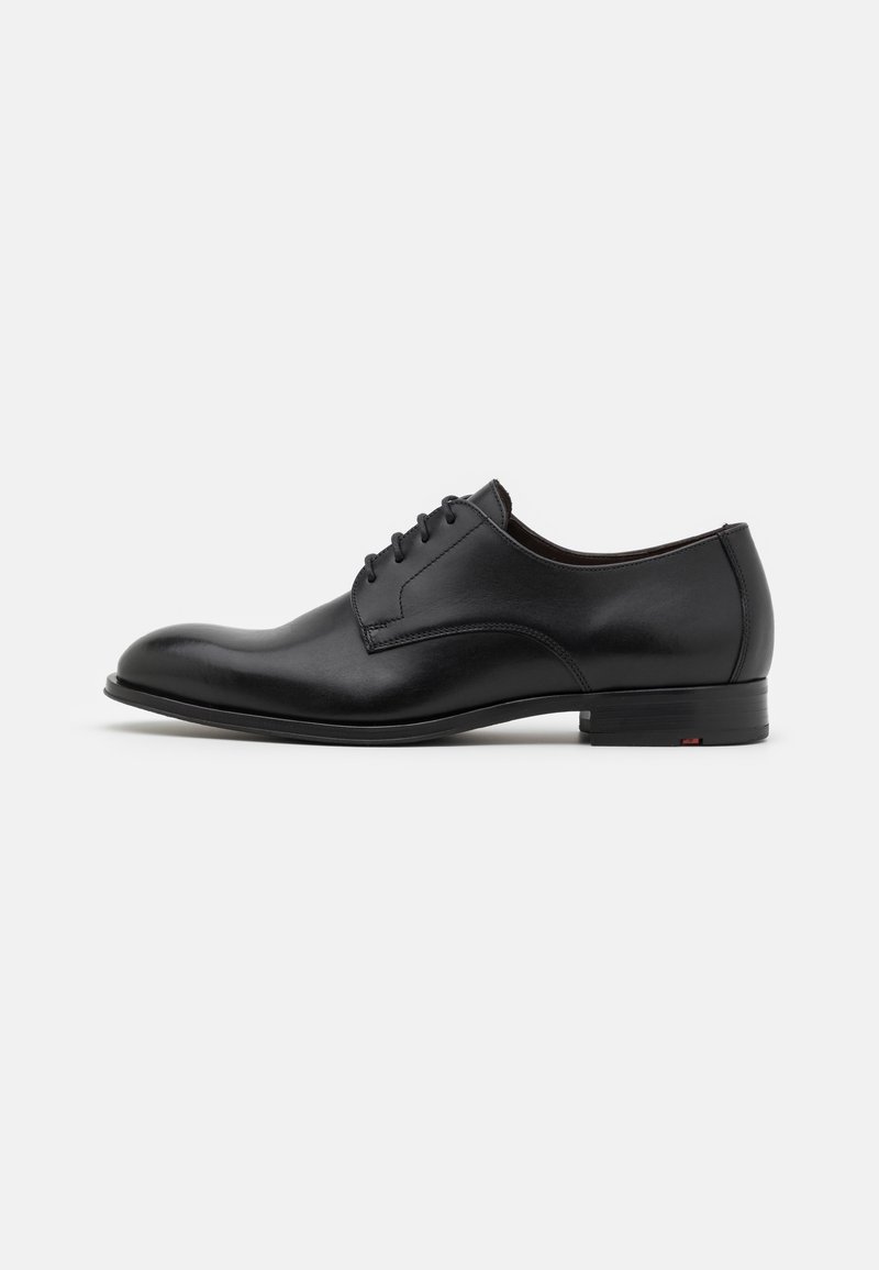 Chaussure de ville en cuir noir avec un bout rond, cinq œillets et un design minimaliste. Elle présente un talon plat et des détails de couture subtils.