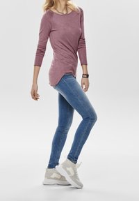 Langärmliges, mauvefarbenes Oberteil mit rundem Ausschnitt, kombiniert mit eng anliegenden blauen Jeans und hellgrauen Sneakers mit einem weißen Akzent.