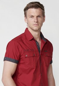 Camisa roja de manga corta con acentos grises, frente abotonado, ajuste entallado y dos bolsillos en el pecho, con costuras decorativas y detalle del logo.