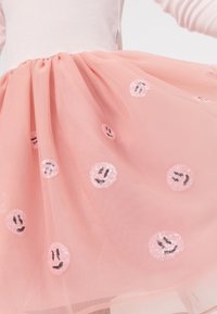Ružová tylová sukňa so sequinmi v tvare usmievajúcej sa tváre, vrstvený dizajn, mäkká textúra a nariasený pás. Akcenty zahŕňajú malé čierne detaily na tvárach.
