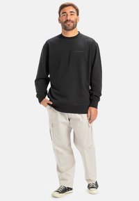 Sudadera negra de cuello redondo con un pequeño logo, combinada con pantalones cargo beige que tienen bolsillos laterales y zapatillas negras de corte bajo.