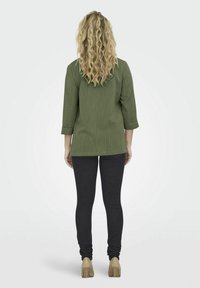 Camisa ligera de color verde oliva con mangas de tres cuartos, combinada con jeans oscuros y zapatos beige, vista desde la parte de atrás. Tejido texturizado.