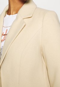 Blazer beige con taglio sartoriale, dotato di colletto a rever, texture liscia e dettagli di cucitura discreti.
