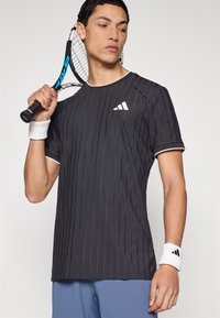 Maglietta atletica grigia a maniche corte con strisce verticali in rilievo, colletto bianco e logo Adidas, indossata con un polsino bianco e una mano che tiene una racchetta.