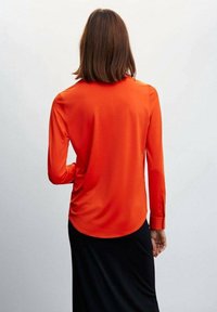 Chemise rouge à manches longues en tissu lisse, avec un poignet boutonné et un ourlet arrondi, portée avec une jupe noire.