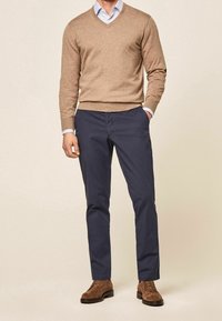 Homme portant un pull beige à col en V, une chemise à col bleu clair, un pantalon bleu marine et des chaussures en daim marron, debout devant un fond uni.