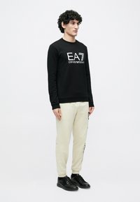 Μαύρο φούτερ με το λογότυπο "EA7 EMPORIO ARMANI", σε συνδυασμό με ανοιχτό μπεζ φόρμα και μαύρα αθλητικά παπούτσια. Το άτομο στέκεται όρθιο.