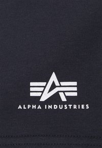 Tamna pamučna tkanina sadrži bijeli logo s geometrijskim dizajnom i tekstom "ALPHA INDUSTRIES", s linijama i emblematskim oblikom.