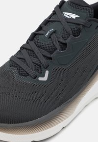 Gray athletic shoe featuring a mesh upper, textured side panels, and a contrasting white and beige sole. Includes black laces and branding.