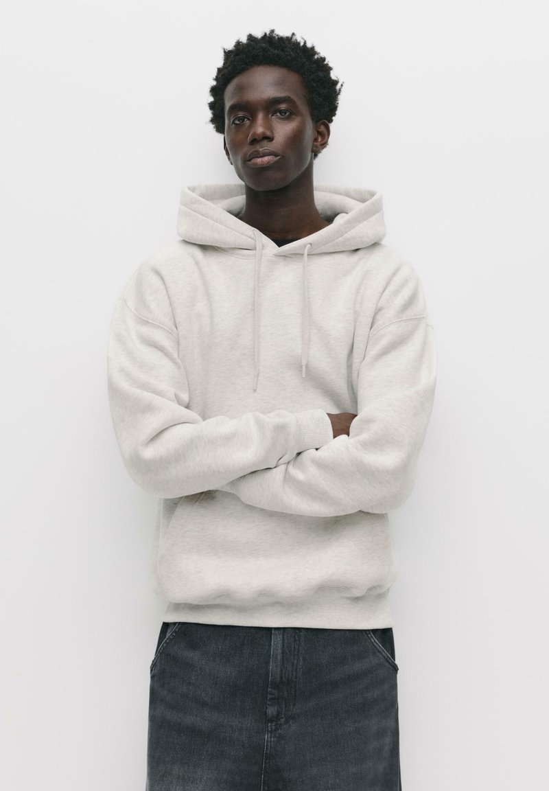 Hellgrauer Kapuzenpullover mit lockerer Passform, gerippten Bündchen und Saum. Getragen mit dunklen Jeans, der Hintergrund ist schlicht und weiß.