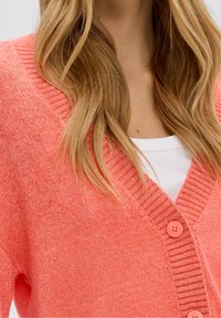 Koralowy sweter na drutach z dekoltem w kształcie litery V, z prążkowanymi detalami wokół kołnierza i trzema różowymi guzikami z przodu. Miękka tekstura.