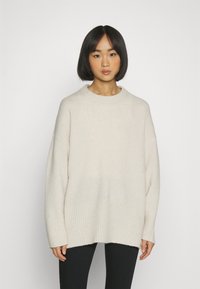 Chunky stickad tröja i ljusbeige, med ribbad nederkant och vida ärmar för en avslappnad passform. Mjuk, texturerad tyg med rundad halsringning.