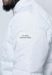 Vit puffjacka med en strukturerad yta, med en rektangulär etikett på vänster axel som säger "LA ROSA COPENHAGEN."