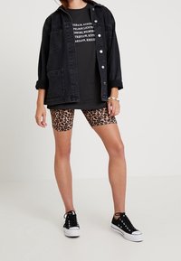 Chaqueta de mezclilla negra sobre una camiseta gráfica negra, combinada con pantalones cortos de ciclismo de estampado de leopardo y zapatillas negras. Incluye bolsillos y botones.