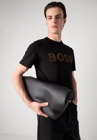 Juodas marškinėliai su ryškiu auksiniu užrašu "BOSS", derinami su tamsiais džinsais. Modelis laiko sulankstytą juodą odinę rankinę.