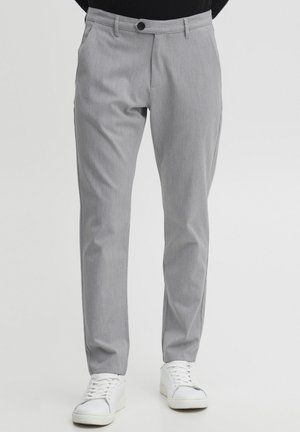 Pantaloni grigi chiari realizzati in tessuto testurizzato, con un design aderente, passanti per la cintura, chiusura frontale con bottone e tasche laterali. Indossati con sneakers bianche.