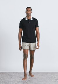 Άνδρας που περπατά ξυπόλητος φορώντας μαύρο πόλο με λευκό γιακά και μπεζ σορτς με σχέδια, πάνω σε απλό φόντο.