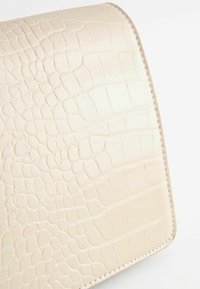 Portfel wytłaczany wzorem krokodyla w jasnobeżowej skórze, o gładkiej fakturze i zaokrąglonych krawędziach, z widocznym przeszyciem wzdłuż szwów.