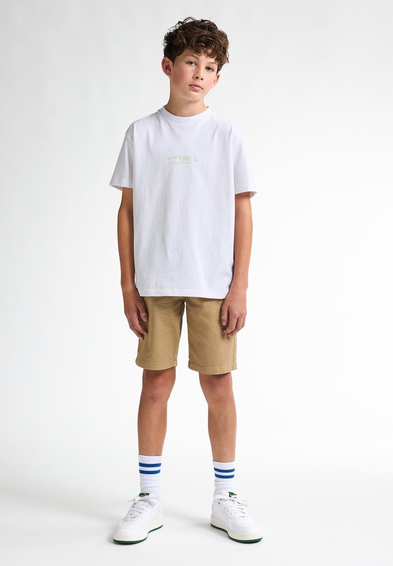 T-shirt bianca in cotone con testo verde, pantaloni shorts khaki, sneaker bianche e calzini bianchi con righe blu. Il modello è in piedi su uno sfondo neutro.