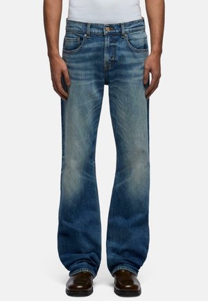 Mand iført falmede blå jeans med lige ben og mørkebrune sko, står med hænderne afslappet ved siden.