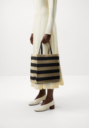 Personne en longue robe crème et chaussures blanches à talons blocs tenant un grand sac fourre-tout tissé rayé noir et beige.