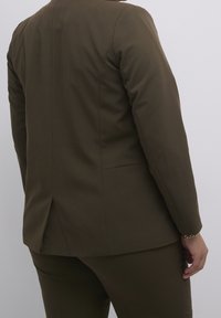 Veste de costume sur mesure marron, texture lisse, une seule fente à l'arrière, manches ajustées, associée à un pantalon assorti, tous deux dans une teinte douce.