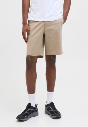 Persona che indossa pantaloncini beige fino al ginocchio, calzini bianchi e scarpe da ginnastica nere, in piedi su uno sfondo semplice.